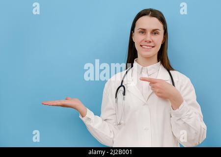 Ritratto di giovane donna felice in professionale medico bianco cappotto indica con mani su spazio copia per la pubblicità. Dottore che presenta e mostra s Foto Stock