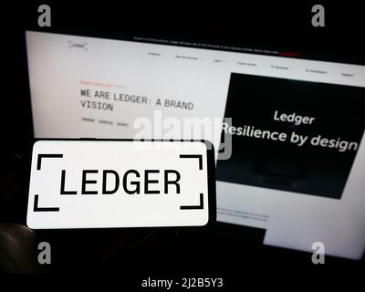 Persona che tiene il cellulare con il logo della società francese di asset digitali Ledger SAS sullo schermo di fronte al sito web aziendale. Mettere a fuoco sul display del telefono. Foto Stock
