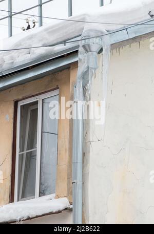 Grandi ghiaccioli appendono dal tubo di drenaggio sul bordo del tetto. Sullo sfondo del muro di una vecchia casa di mattoni. Grandi cascate, anche belle file. Nuvoloso giorno d'inverno, luce tenue. Foto Stock