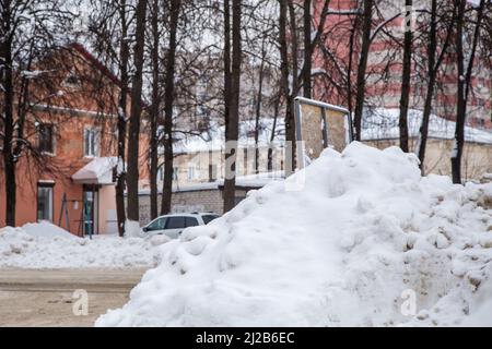 La neve sporca deriva dalla strada contro lo sfondo delle case e degli alberi della città. Sulla strada si trova neve sporca in cumuli alti. Paesaggio urbano invernale. Nuvoloso giorno d'inverno, luce tenue. Foto Stock