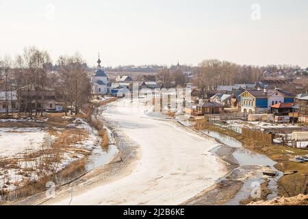 Un fiume storto e ghiacciato che si estende in lontananza, case di legno sulla riva. Primavera, neve si scioglie, pozzanghere e erba secca tutto intorno. Giorno, tempo nuvoloso, luce calda e soffusa. Foto Stock