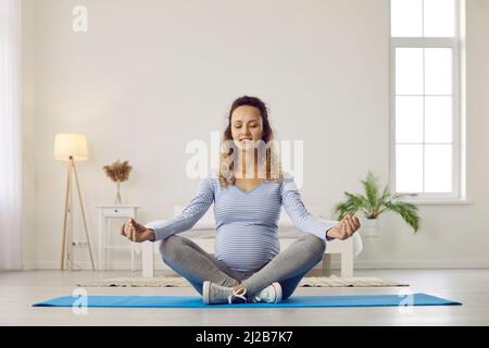 Donna incinta calma medita seduta in posizione padmasana yoga sul pavimento a casa. Foto Stock