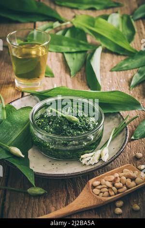 Pesto all'aglio selvatico fatto in casa in una ciotola di vetro su sfondo di legno, decorato con foglie, parmigiano e olio d'oliva, colori desaturati, verticale Foto Stock