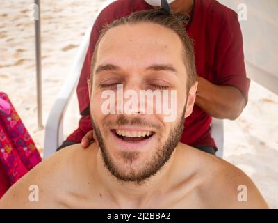L'uomo bianco con gli occhi chiusi sta godendo il massaggio della testa che sta ottenendo alla spiaggia Foto Stock