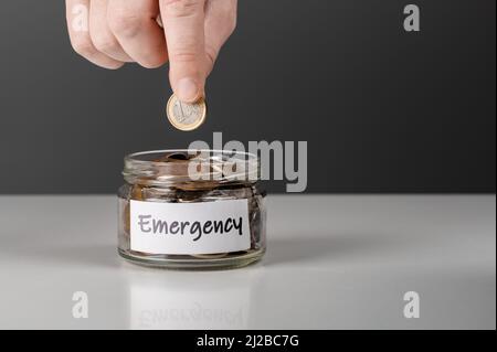 Mano uomo mettere denaro in vaso con testo di emergenza. Mano mette una moneta euro in vaso con la parola emergenza. Mano mettere moneta nel vaso di vetro per savi Foto Stock