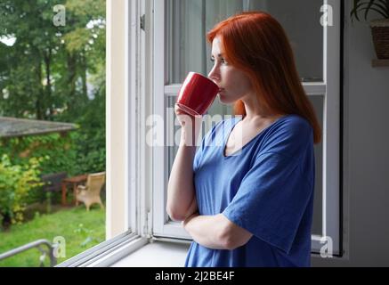 giovane donna che guarda fuori dalla finestra mentre beve dalla tazza di caffè Foto Stock