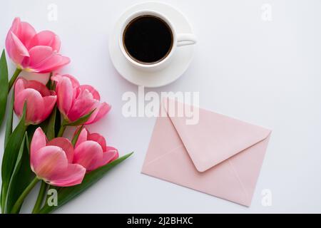 vista dall'alto del bouquet di tulipani vicino a una tazza di caffè e busta rosa su bianco Foto Stock