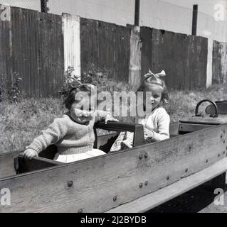 1961, fuori in un parco giochi, due bambine che siedono in un cavallo dondolo incorniciato in legno, Inghilterra, Regno Unito. Il primo parco giochi al mondo risale alla metà del 1800s, dove è stata creata un'area per i bambini con blocchi di legno e giocattoli. Il parco giochi, come lo conosciamo oggi, si è sviluppato come un modo per incoraggiare i bambini fuori dalle strade e dal 1930s in poi, strutture in legno e metallo, come scivoli, altalene, cornici da arrampicata e giostre, ha fornito un punto di partenza per i bambini per divertirsi all'aperto. Con l'introduzione di nuove norme in materia di salute e sicurezza, tali strutture sono state eliminate dal 1980s. Foto Stock