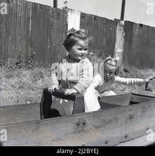 1961, fuori in un parco giochi, due bambine che siedono in un cavallo dondolo incorniciato in legno, Inghilterra, Regno Unito. Il primo parco giochi al mondo risale alla metà del 1800s, dove è stata creata un'area per i bambini con blocchi di legno e giocattoli. Il parco giochi, come lo conosciamo oggi, si è sviluppato come un modo per incoraggiare i bambini fuori dalle strade e dal 1930s in poi, strutture in legno e metallo, come scivoli, altalene, cornici da arrampicata e giostre, ha fornito un punto di partenza per i bambini per divertirsi all'aperto. Con l'introduzione di nuove norme in materia di salute e sicurezza, tali strutture sono state eliminate dal 1980s. Foto Stock