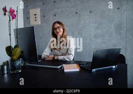 Una donna anziana ben vestita con occhiali moderni si siede alla sua scrivania in ufficio con entrambe le mani sulla scrivania. Guarda la fotocamera. Foto Stock