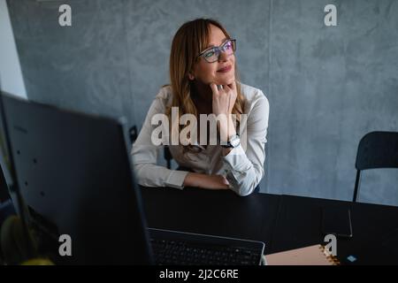 Bella donna caucasica anziana con occhiali si siede alla sua scrivania in ufficio e guarda pensosamente fuori dalla finestra. Si posò il mento sulla mano sinistra. Foto Stock
