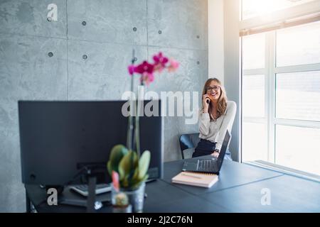 Donna caucasica di mezza età sorridente con occhiali, seduta su una sedia, accanto alla finestra nell'angolo dell'ufficio, fare una telefonata. Vaso di fiore rosa. Foto Stock