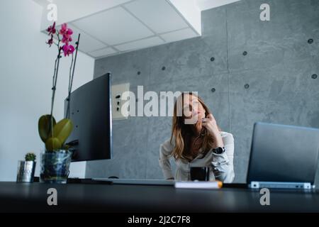 La donna più anziana con occhi verdi immaginata guarda con attenzione verso l'esterno la window.Her mano destra poggia sul suo lato, mentre nella sua mano sinistra, lei tiene gli occhiali. Foto Stock
