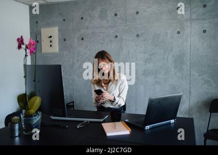 Una bella donna digita messaggi sul suo smartphone mentre si siede di fronte alla sua scrivania. C'è un laptop, un desktop, un notebook, occhiali e penna. Foto Stock