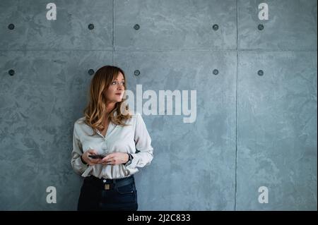 Moderna, bella donna caucasica di mezza età manager sta guardando attraverso la finestra e tenendo il suo smartphone.She è appoggiata su uno sfondo grigio. Foto Stock