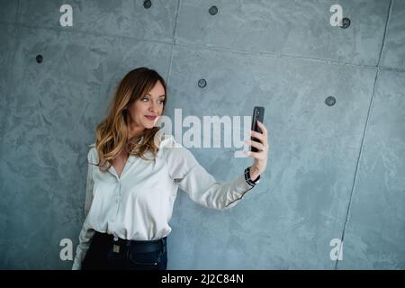 Un manager felice e soddisfatto sta guardando la fotocamera del suo smartphone durante il periodo di pausa sul luogo di lavoro in azienda. Donna anziana caucasica. Foto Stock