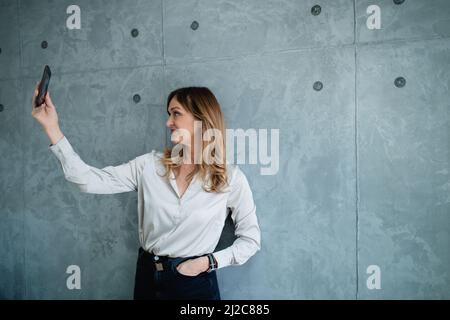 Una donna di mezza età dai capelli castani in una camicia bianca sta facendo selfie con il suo smartphone durante il suo lavoro nel suo moderno ufficio. Foto Stock