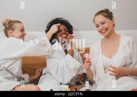 Le giovani donne in accappatoi sono sedute sul letto e utilizzando prodotti per la cura della pelle Foto Stock