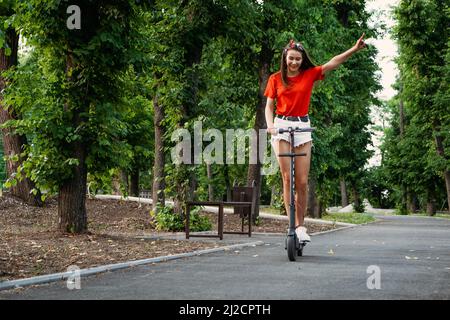E-bike, scooter sicurezza. Giovane donna che guida e scooter in ambiente urbano. Donna alla moda in scooter elettrico nel parco cittadino Foto Stock