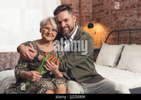 Nonna caucasica soddisfare in un abbraccio con il suo nipote medio-vecchio uomo d'affari che celebra il giorno del nonno. Foto di alta qualità Foto Stock