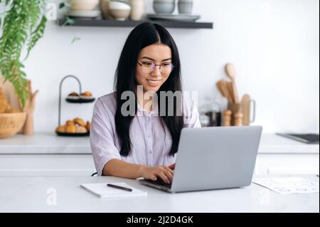 Bella positiva ragazza asiatica intelligente con occhiali, freelancer o studente, si siede a casa in cucina, utilizza un computer portatile per il lavoro remoto o lo studio, naviga in Internet, notizie, sorrisi Foto Stock