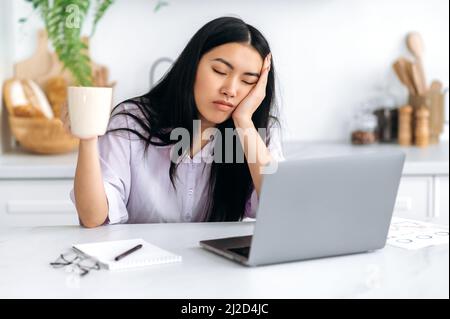 Una ragazza asiatica assonnata, una freelancer o uno studente, si siede a casa in cucina a un tavolo con gli occhi chiusi di fronte a un computer portatile, tiene in mano una tazza di caffè, non può svegliarsi e iniziare a lavorare Foto Stock