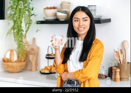 Stile di vita sano. Presa d'acqua giornaliera. Positiva bella ragazza cinese in abiti casual elegante, in piedi a casa in cucina, tenendo un bicchiere di acqua pulita, guardando via, sognando, sorridendo Foto Stock