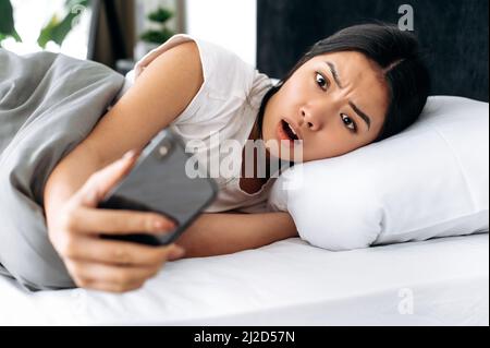 Scioccata sorpresa ragazza asiatica sdraiata in un letto al mattino, guarda spaventato al suo smartphone, ha dormito la sveglia, emotiva espressione facciale, emozione negativa, notizie inaspettate Foto Stock