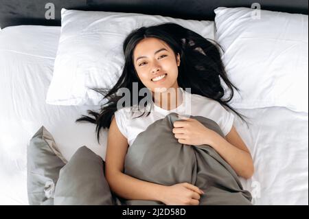 Vista dall'alto di una bella ragazza bruna asiatica positiva in una t-shirt bianca, sdraiata sotto le coperte a letto a casa al mattino, guarda la macchina fotografica, sorride, si rilassa in un letto accogliente durante il fine settimana Foto Stock