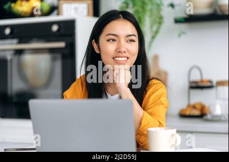 Bella bruna asiatica positiva giovane donna, designer, freelancer o studente, si siede in cucina con un laptop, lavorando o studiare a distanza, guardando al lato, pensando, sognando, sorride amichevole Foto Stock