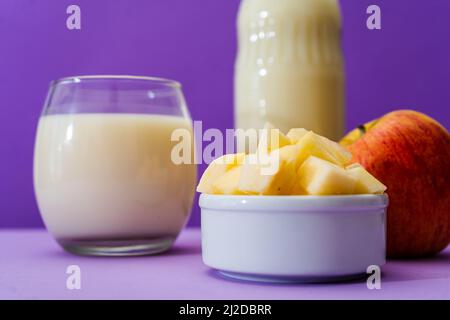 Bottiglia di vetro e bicchiere grande con latte e una mela rossa intera accanto a una ciotola con cubetti di mela in ambiente viola o viola. Spazio di copia. Foto Stock