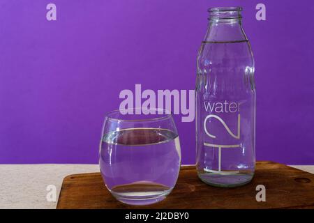 Bicchiere di vetro e bottiglia riempiti con acqua purificata. Sfondo viola. Foto Stock