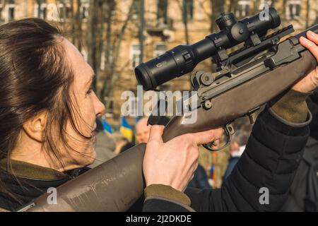 Bella ragazza o donna ucraina con un fucile o fucile di addestramento per la guerra o la battaglia in città, con bandiere ucraine sullo sfondo Foto Stock