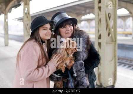 Mamma e figlia si sono incontrate con gioia sulla piattaforma di stazione e abbracciano, in mani piccolo cane, c'è una grande valigia a terra nella stagione invernale nevoso. Rapporto in famiglia Foto Stock