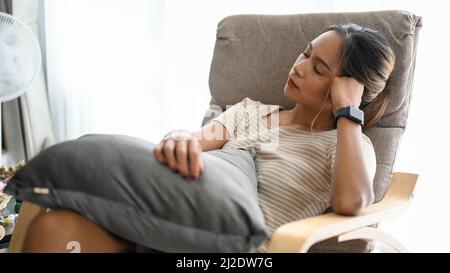 Una bella donna asiatica che fa un pisolino su una comoda poltrona nel suo luminoso soggiorno. Riposo e relax Foto Stock