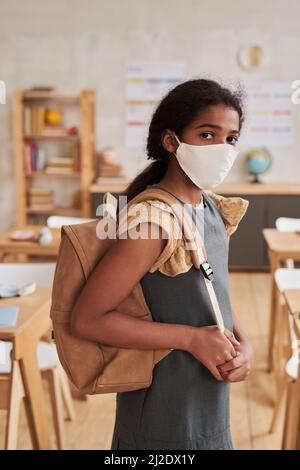 Ritratto verticale della ragazza afro-americana che indossa la maschera a scuola e guarda la macchina fotografica mentre si trova in aula Foto Stock