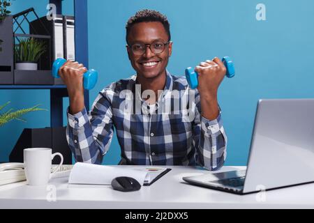 Uomo d'affari atletico azienda che solleva pesi mentre si siede alla scrivania in ufficio. Giovane adulto attivo e sano che fa l'allenamento con manubri fitness al fine di mantenere il corpo forte. Foto Stock