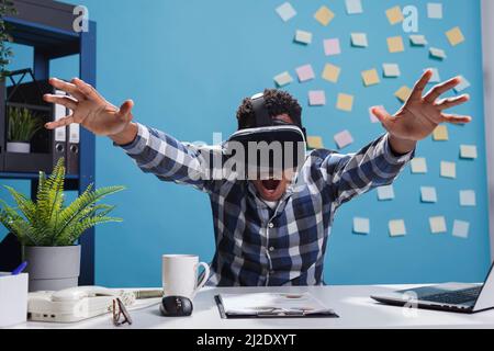 L'espressivo stupito impiegato dell'azienda afroamericana che gode della tecnologia di gioco di realtà virtuale. Uomo d'affari adulto che indossa un dispositivo di intrattenimento elettronico mentre si siede alla scrivania nello spazio di lavoro. Foto Stock