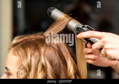 Parrucchiere ricci con un ferro da stiro per la giovane donna con lunghi capelli marroni in un salone di bellezza Foto Stock