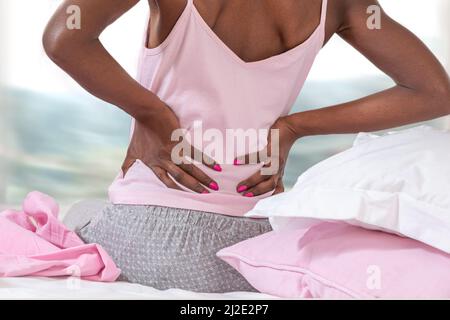 Vista posteriore di una giovane donna africana e americana seduta a letto e affetta da mal di schiena Foto Stock