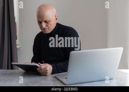 Confuso grigio capelli calvo 60 anni maschio in abiti formali di navigazione moderno tablet con bewilderment seduta in poltrona al tavolo Foto Stock