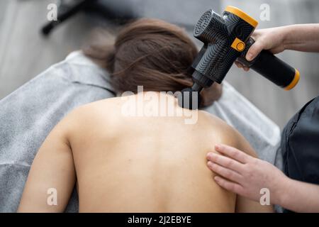 La donna massaggia la schiena del paziente con un massaggiatore a percussione Foto Stock