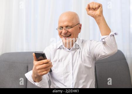 Eccitato anziano anziano pensionato uomo che guarda lo schermo del telefono, che celebra la vittoria in linea della lotteria o che ottiene il messaggio con le buone notizie. Emotivo maturo Foto Stock