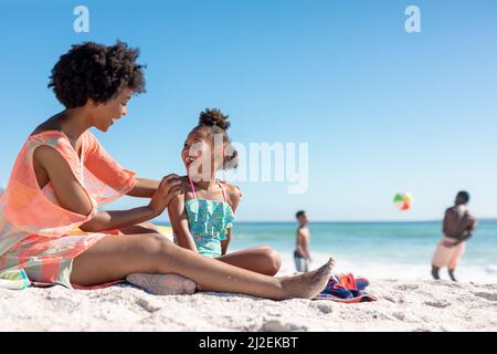 Allegra madre e figlia afroamericana seduta insieme mentre padre e figlio giocano in spiaggia Foto Stock