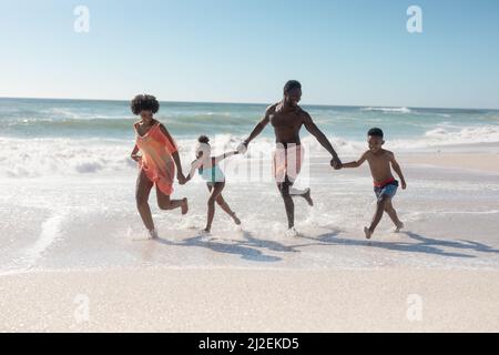 Famiglia afro-americana che tiene le mani mentre corre insieme in spiaggia in giorno di sole Foto Stock