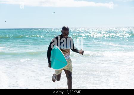 Felice uomo maggiore afroamericano che trasporta la tavola da surf che corre sulla riva alla spiaggia contro il cielo Foto Stock