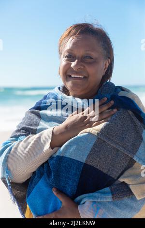 Sorridente donna anziana in pensione afroamericana avvolta in scialle guardando via alla spiaggia in giorno di sole Foto Stock