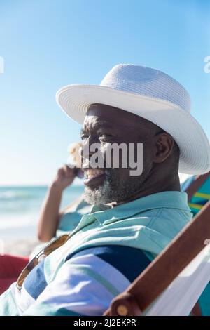 Uomo anziano afroamericano che indossa un cappello seduto su sedia con una donna in spiaggia durante la giornata di sole Foto Stock