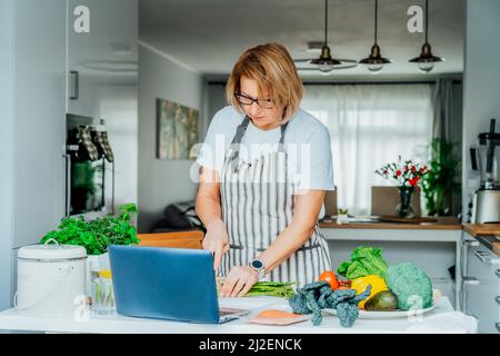 Donna di mezza età seguendo un tutorial di cucina video corso sul portatile mentre si prepara il pasto in una cucina. Donna che cucina sano piatto, pesce e verdure Foto Stock