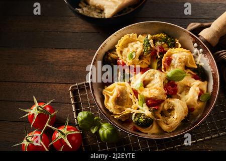 I tortelloni italiani con broccoli e pomodori in padella calda su griglia metallica vicino al parmigiano su tavola di legno Foto Stock